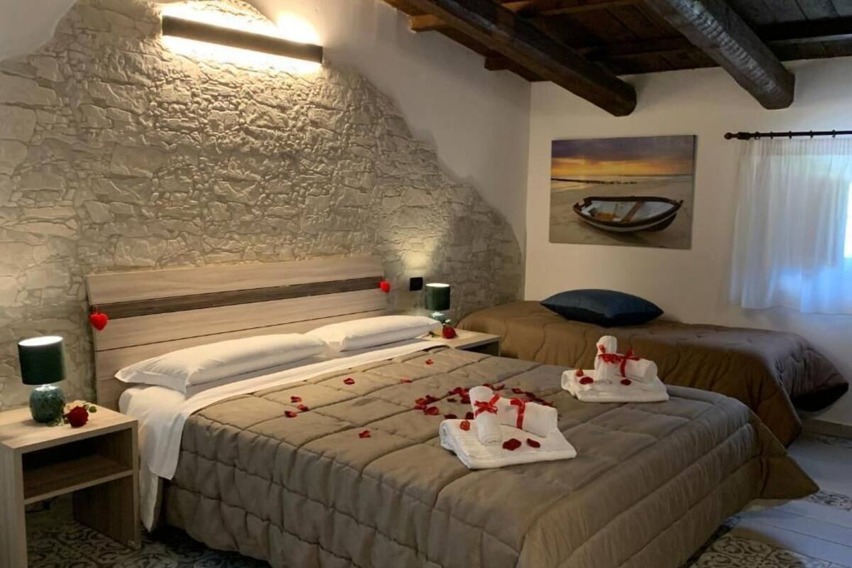 A bedroom with a stone wall, a bed with rose petals, a painting of a boat, and a window with curtains.