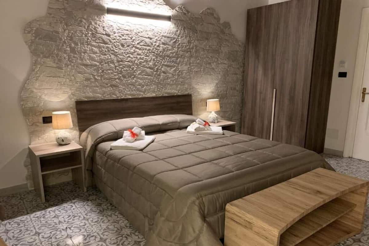 A bedroom with a stone wall, a bed with a wooden headboard, two bedside tables with lamps, and a wooden bench.