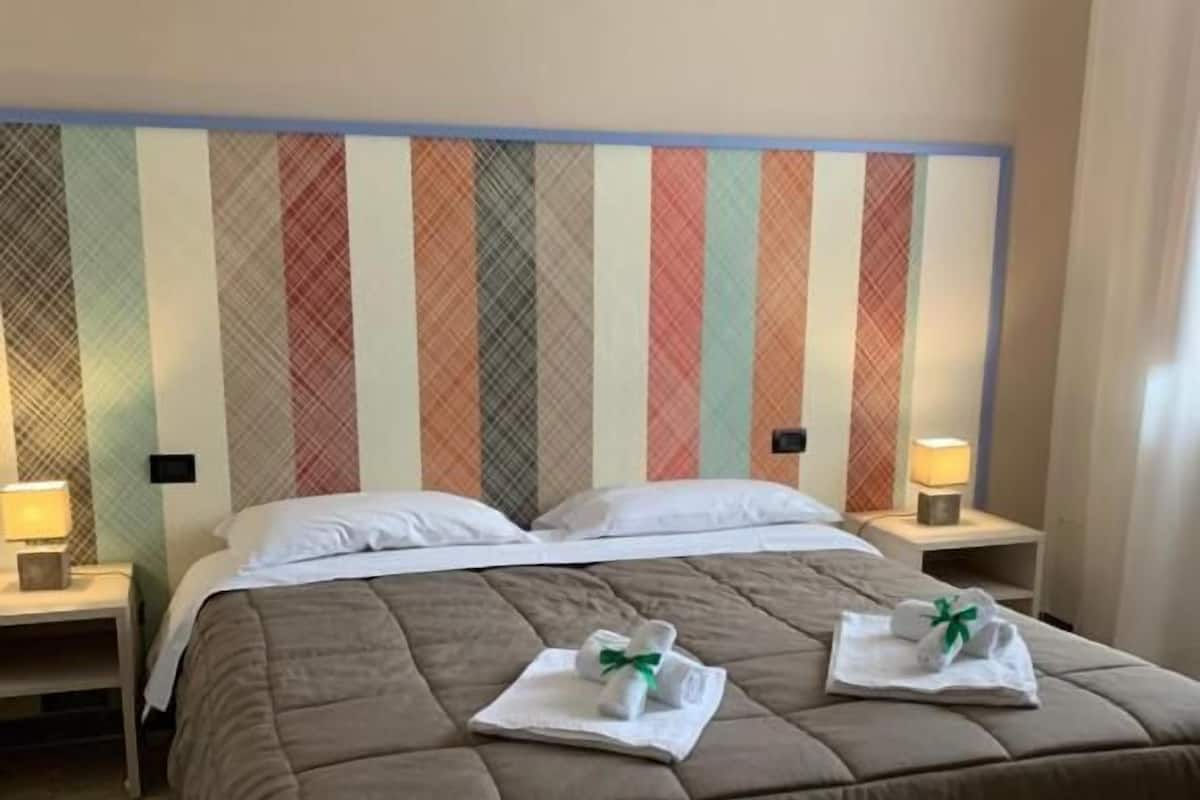 A hotel room with a large bed, two bedside tables with lamps, a wooden nightstand, and a wooden bench.