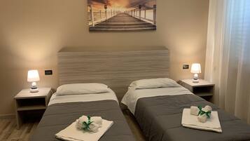 Ein Hotelzimmer mit zwei Betten, jeweils mit Kissen und Handtuch. Nachttische mit Lampen und ein Gemälde von einem Steg an der Wand.