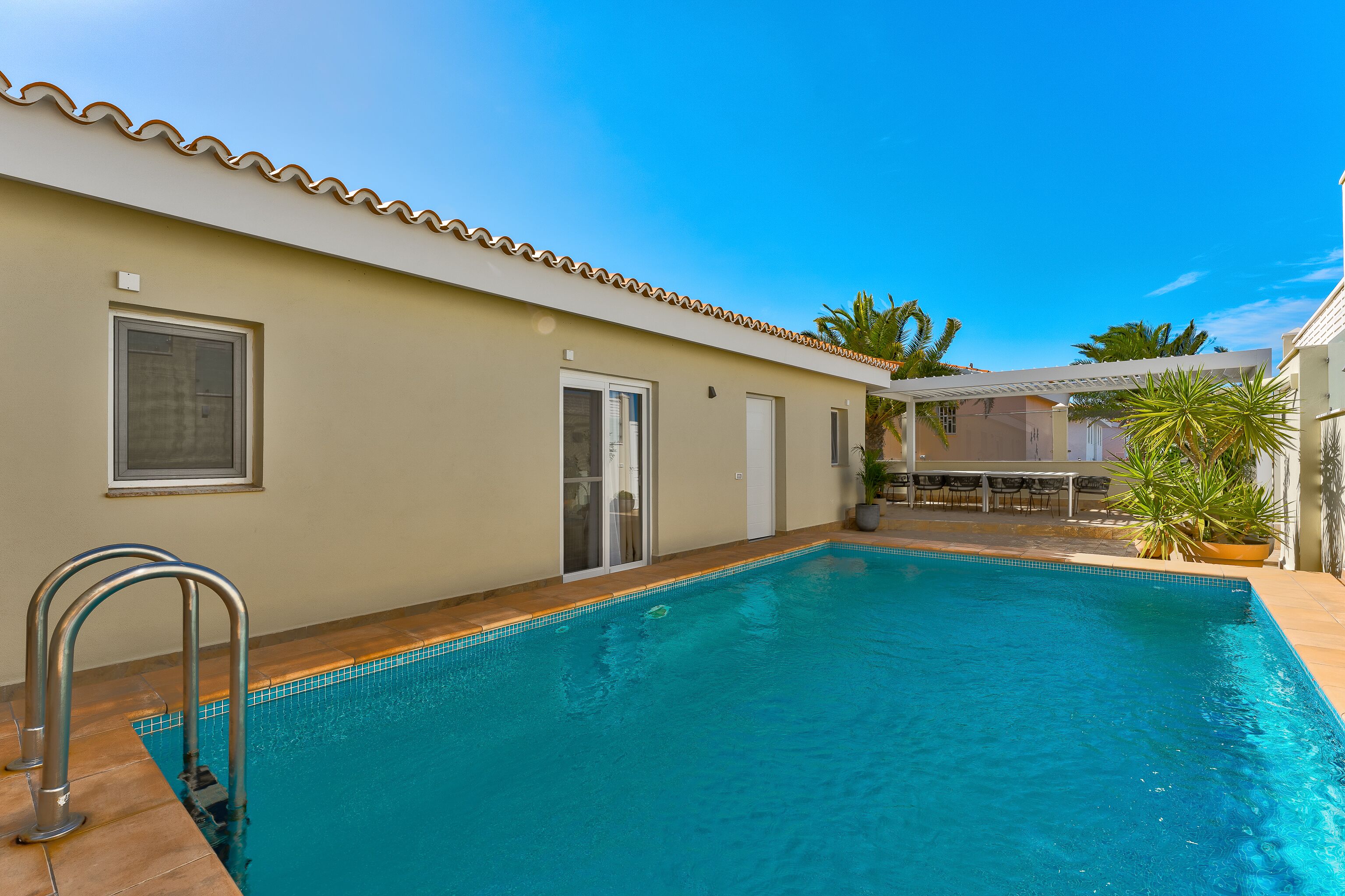 A swimming pool with a tiled edge, a metal ladder, and a door leading to a covered patio area.