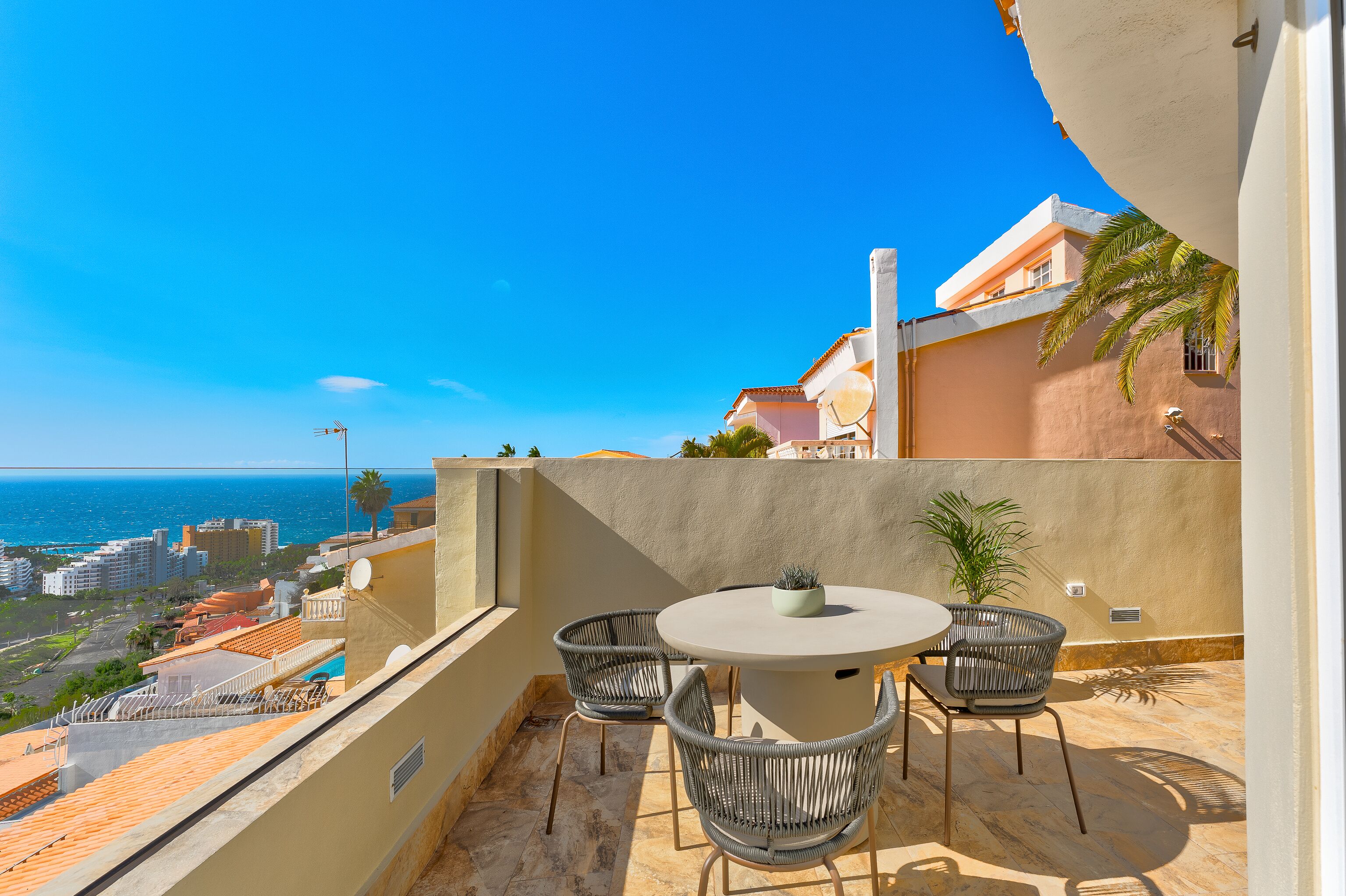 Une terrasse avec une table ronde et des chaises, un pot de fleurs et une vue sur la mer et le paysage urbain.