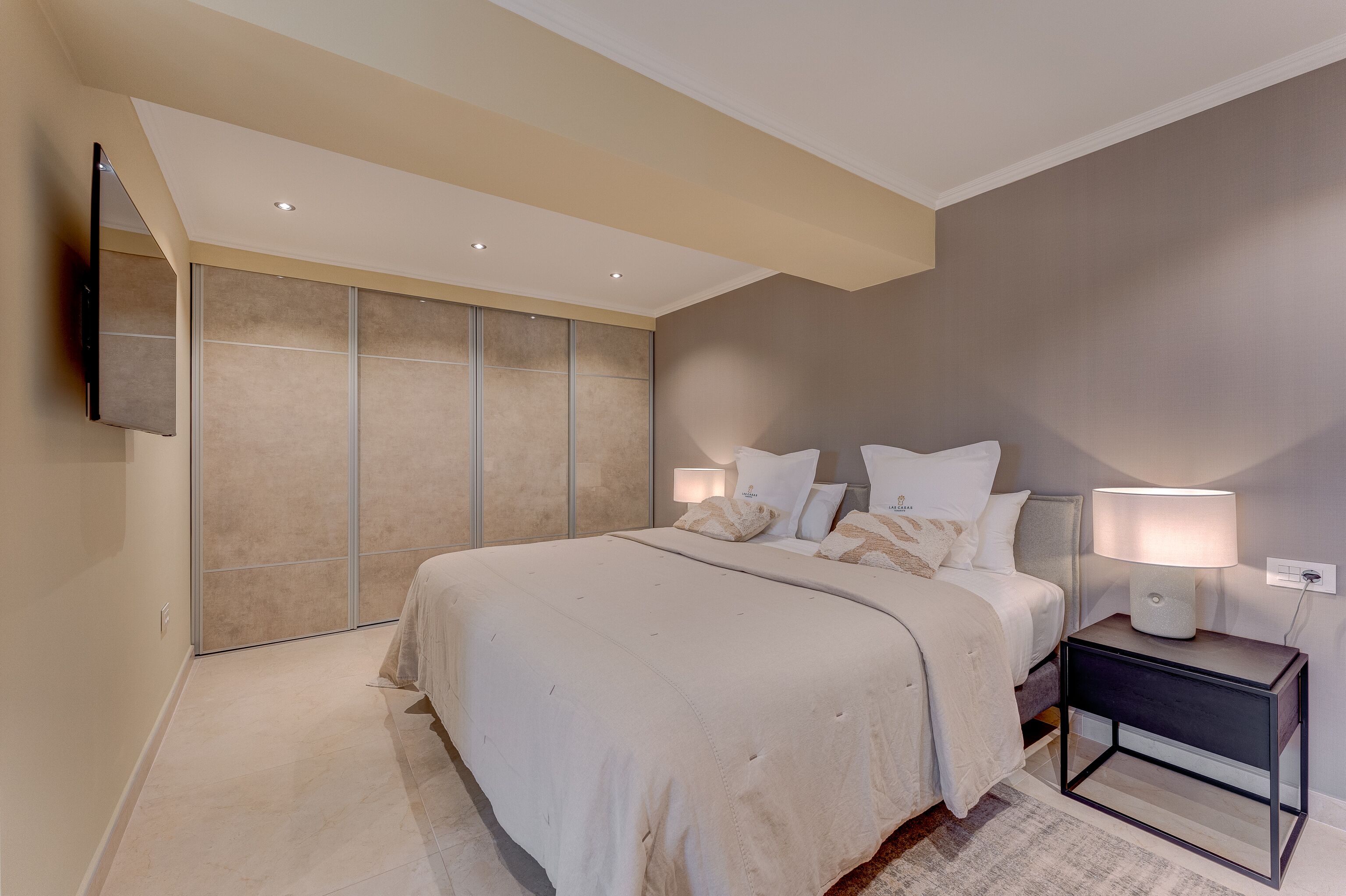 A bedroom with a large bed, a nightstand with a lamp, and a wall-mounted TV.