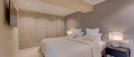 A bedroom with a large bed, a nightstand with a lamp, and a wall-mounted TV.