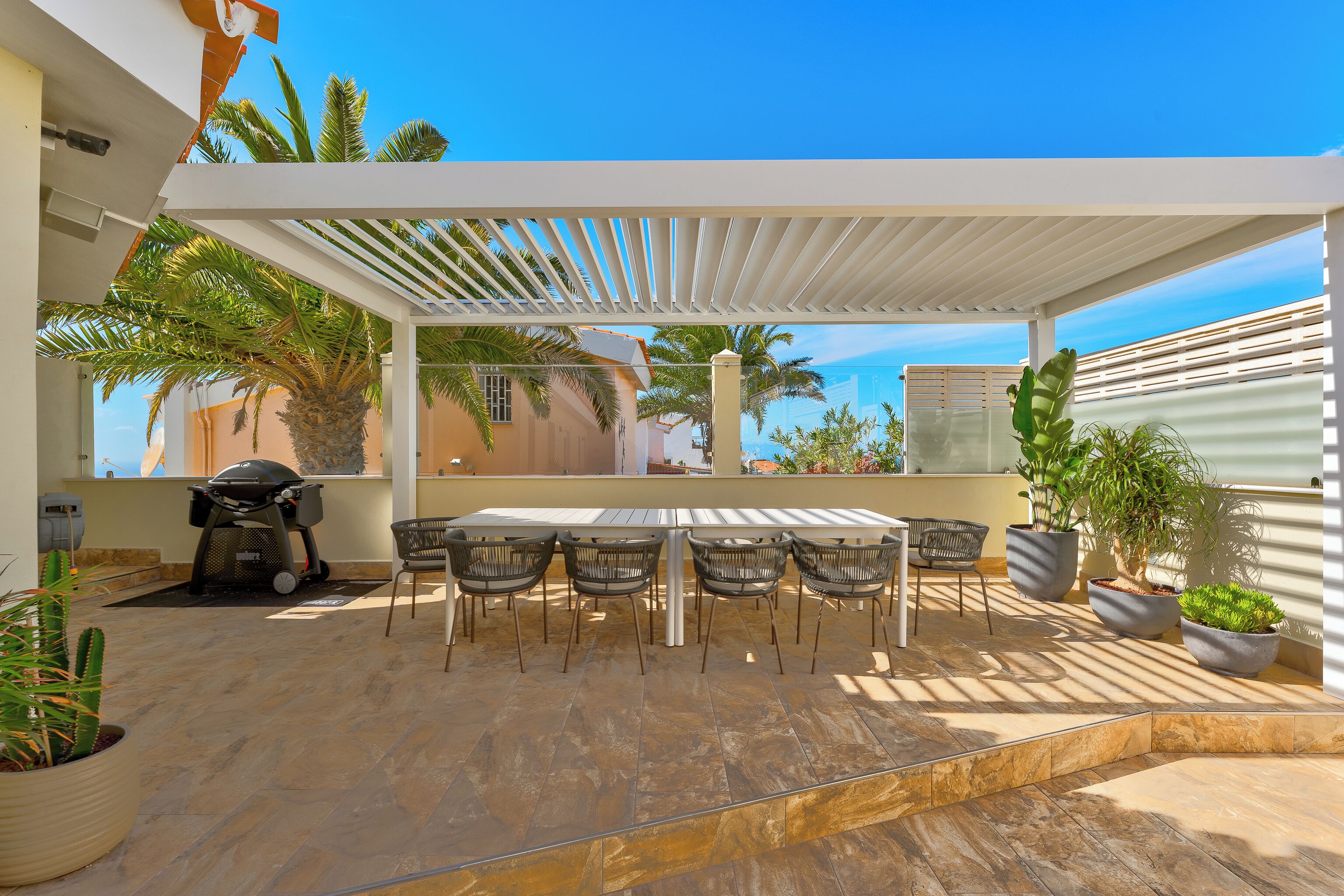 An outdoor patio with a white pergola, a barbecue grill, and a dining table with chairs.