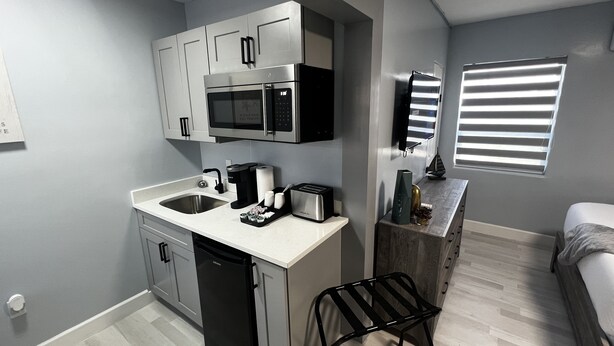 A compact kitchen with a microwave, toaster, and sink. A small dining area with a chair and a window with blinds.