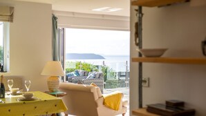 A living room with a sofa, a yellow table, and a lamp. There is a balcony with outdoor furniture and a view of the sea.