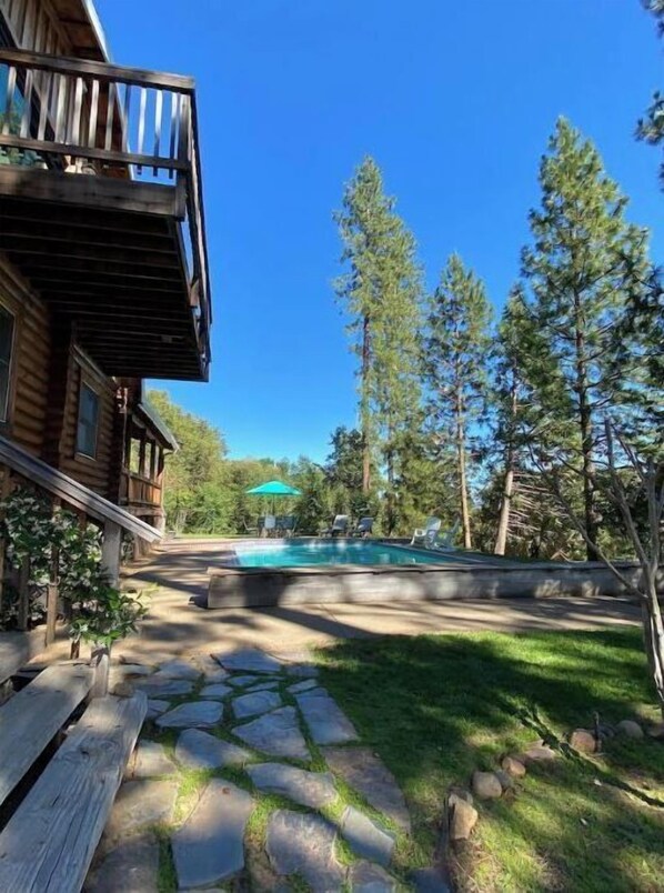 A wooden cabin with a stone pathway leading to a pool area surrounded by tall trees.