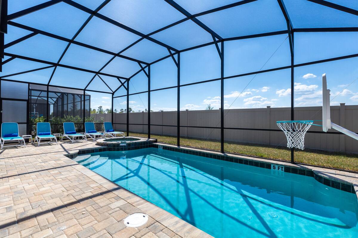 A covered outdoor swimming pool with blue water, a basketball hoop, and lounge chairs.