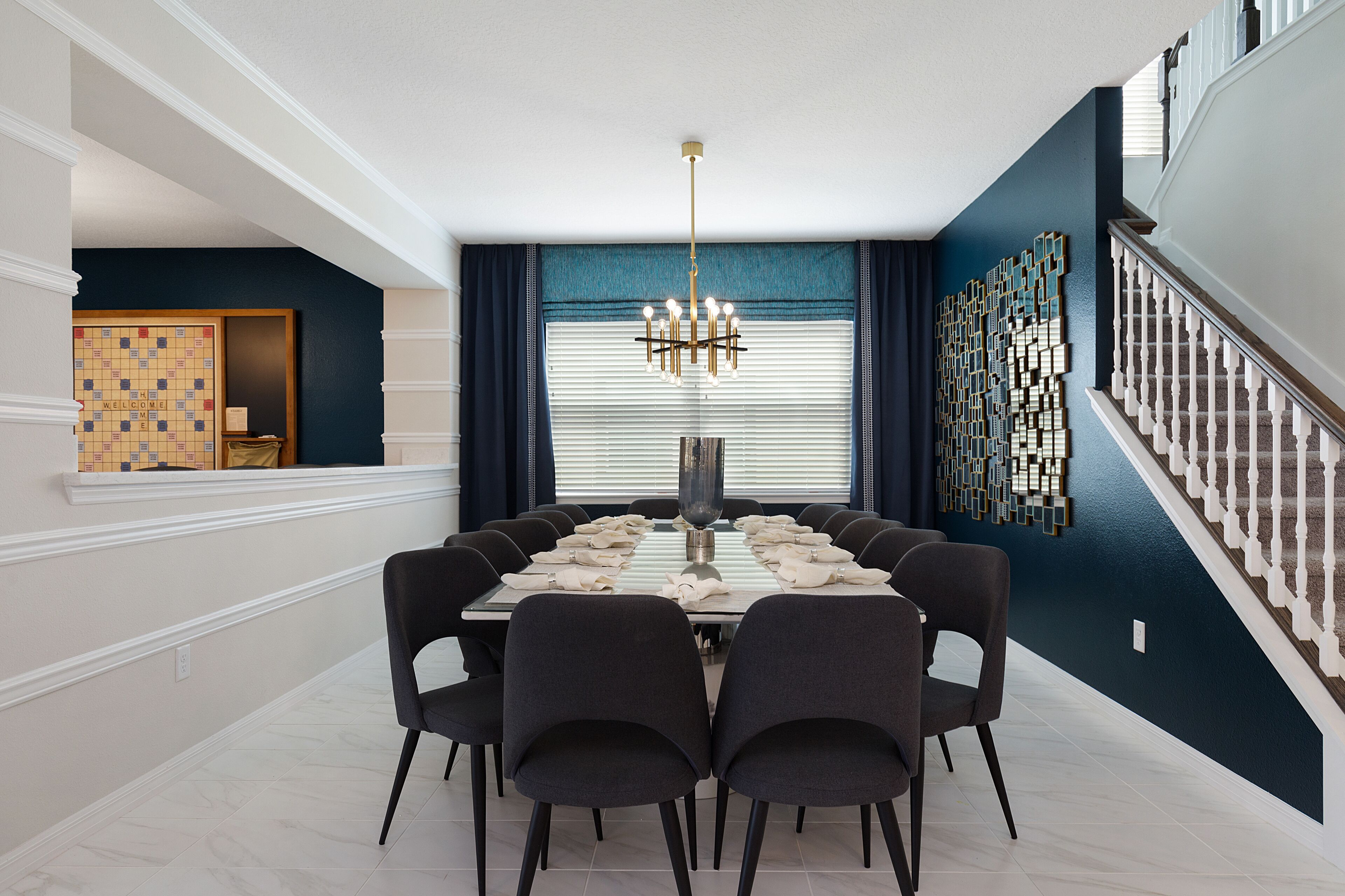 Une salle à manger avec une grande table rectangulaire, des chaises noires, un lustre et un escalier avec des rampes blanches.