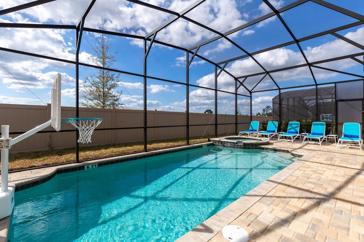 A clear blue pool with a basketball hoop, surrounded by lounge chairs and a covered area with a view of the sky.