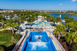 A water park with a large pool, palm trees, and a view of a lake and houses in the background. - Villatel at Solara Resort - 8BRs (Kissimmee)