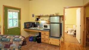 A compact kitchen with a refrigerator, microwave, and sink. There is a window with a curtain, a bed with a patterned blanket, and a door leading to another room. - small 2-bedroom chalet in the heart of the wilderness, 5 km from Kejimkujik Park (Maitland Bridge)