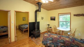 A cozy room with a wood-burning stove, a bed, a table with chairs, and a window with a view of trees. - small 2-bedroom chalet in the heart of the wilderness, 5 km from Kejimkujik Park (Maitland Bridge)