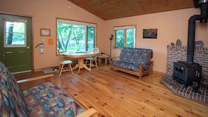 Ein gemütliches Wohnzimmer mit Kamin, Holzboden, einer grünen Tür und großen Fenstern mit Blick ins Freie.