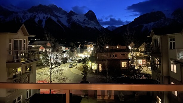 A nighttime view of a mountain town with illuminated buildings and a snow-covered mountain range in the background. - Stunning Mountain Views-Cozy 2 Bedroom (Canmore)