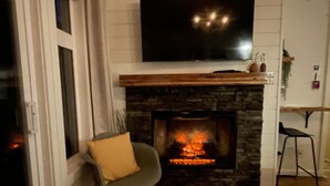 A living room with a stone fireplace, a flat-screen TV mounted above it, and a grey chair with a yellow cushion. - Stunning Mountain Views-Cozy 2 Bedroom (Canmore)