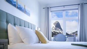 A bedroom with a bed, pillows, a chair, a window with a mountain view, and a painting on the wall. - Stunning Mountain Views-Cozy 2 Bedroom (Canmore)