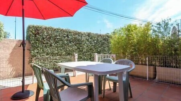 Un patio al aire libre con una mesa, sillas, un paraguas rojo y un seto al fondo.