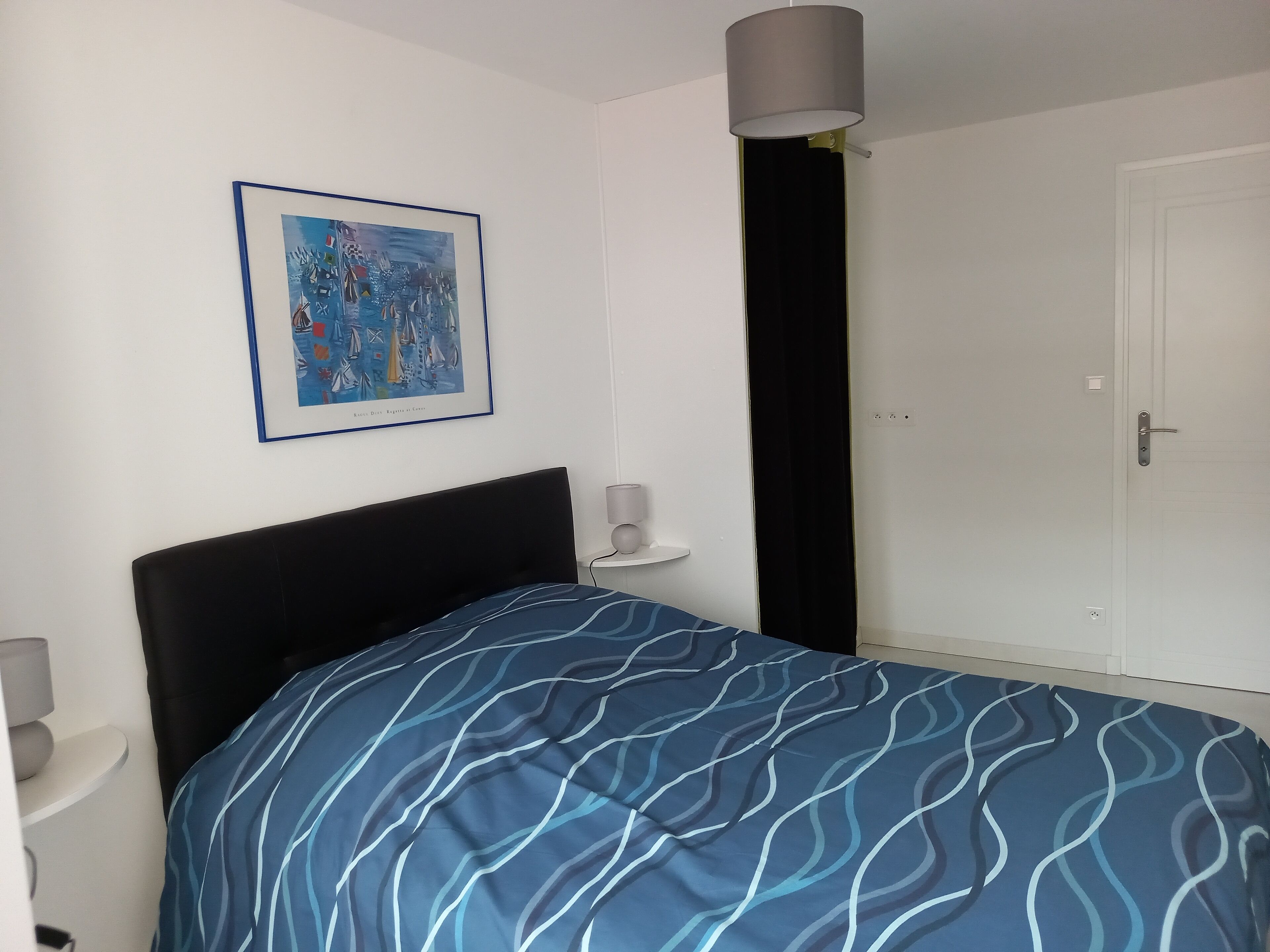 Ein Schlafzimmer mit einem Bett, einem gerahmten Bild an der Wand, einem Nachttisch mit einer Lampe und einer Tür.