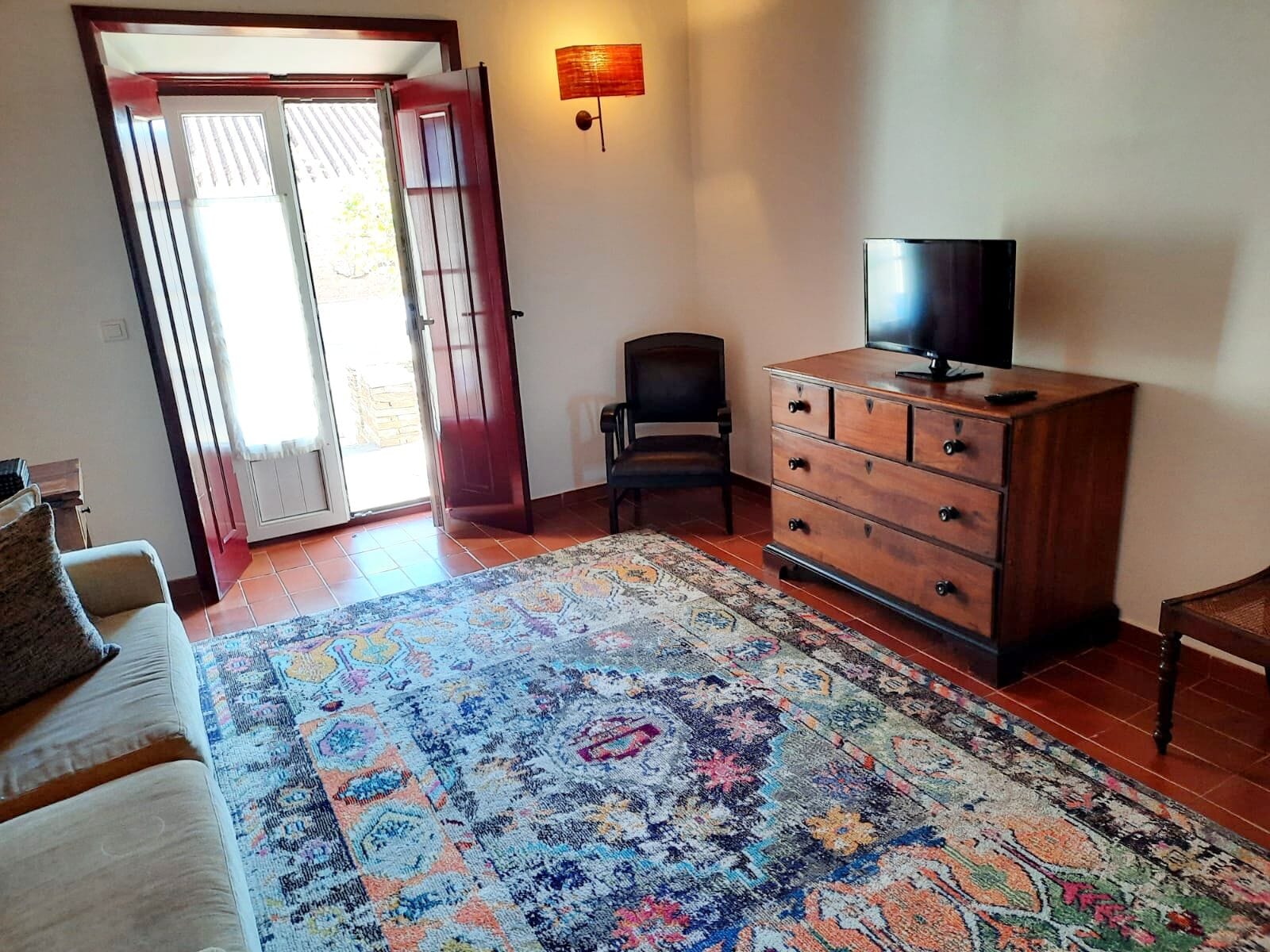 A room with a large patterned rug, a wooden dresser with a TV, a chair, and a sofa.