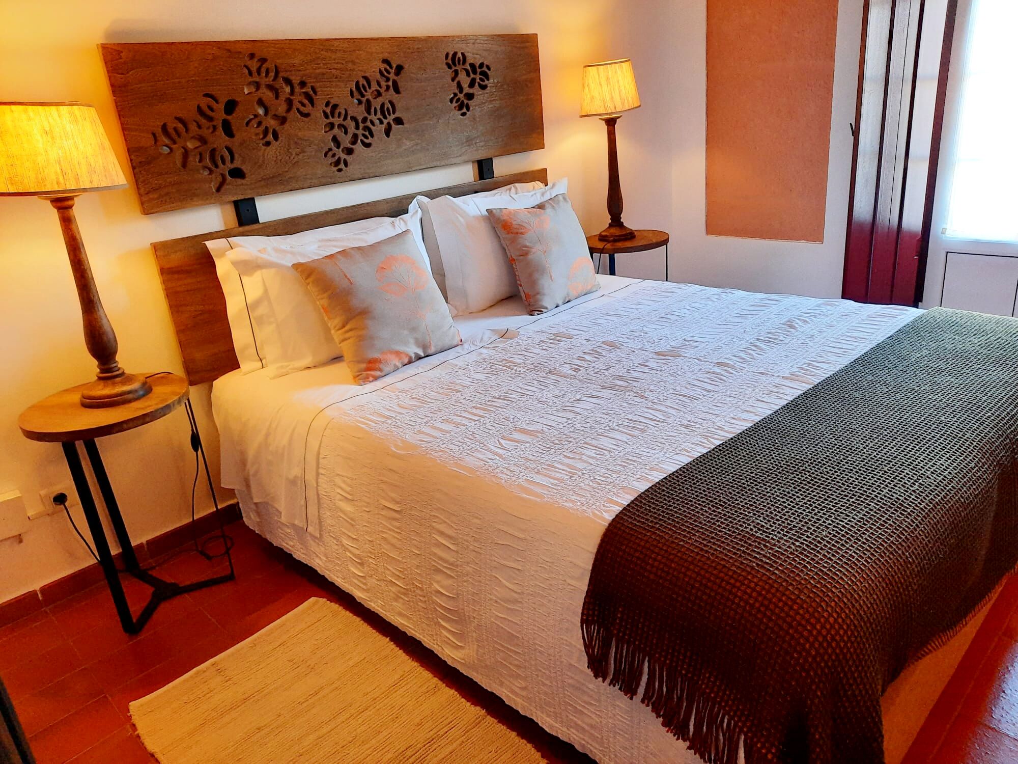 Una camera d'albergo con un letto grande, due lampade da comodino, una testiera decorativa e una finestra con tende a rullo verticali.