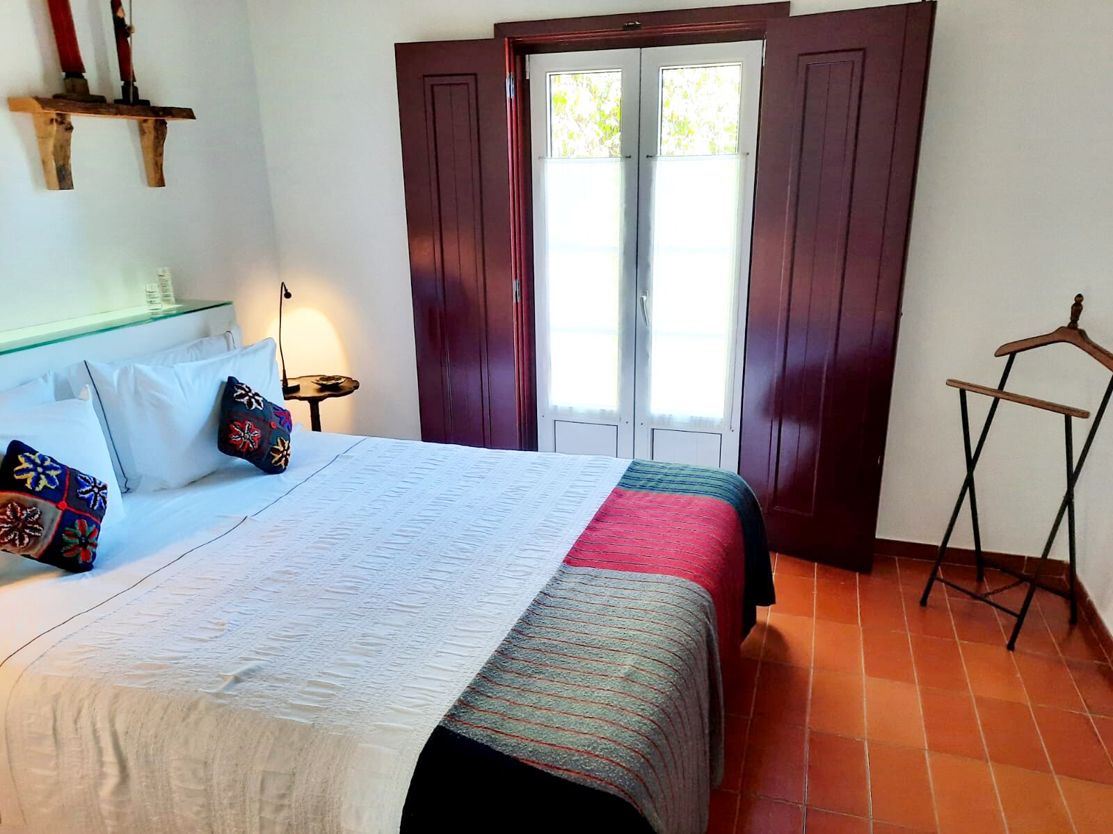 Una cama con un edredón blanco y una manta colorida, un perchero de madera, una mesita con una lámpara y una ventana con contraventanas de madera.
