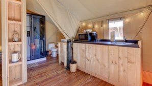 Een compacte keuken met houten kastjes, een magnetron en een spoelbak. Er is een klein raam met lichtslingers en een deur naar een badkamer.
