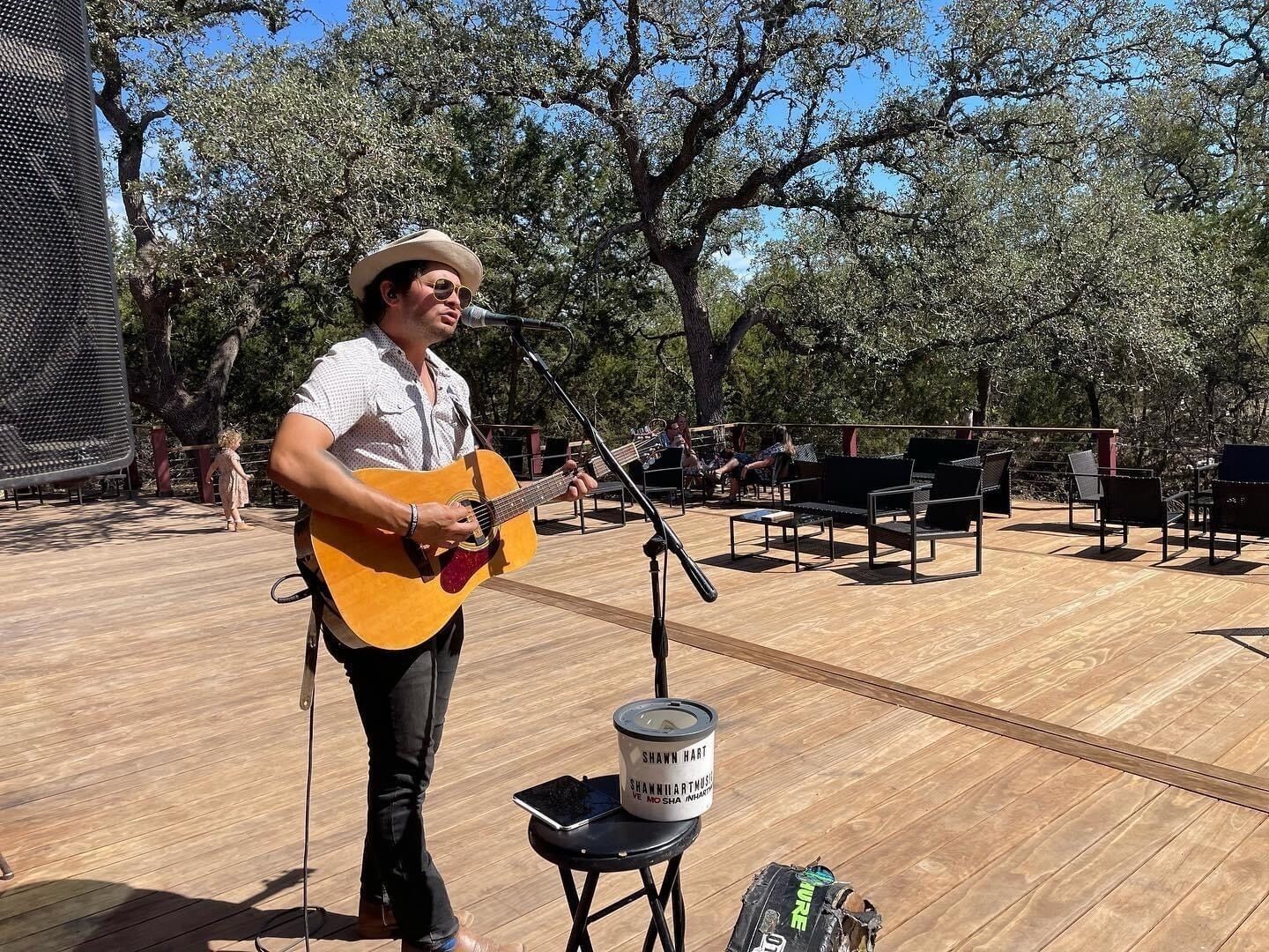 A person playing an acoustic guitar on an outdoor stage with a microphone stand, surrounded by trees and outdoor seating.
