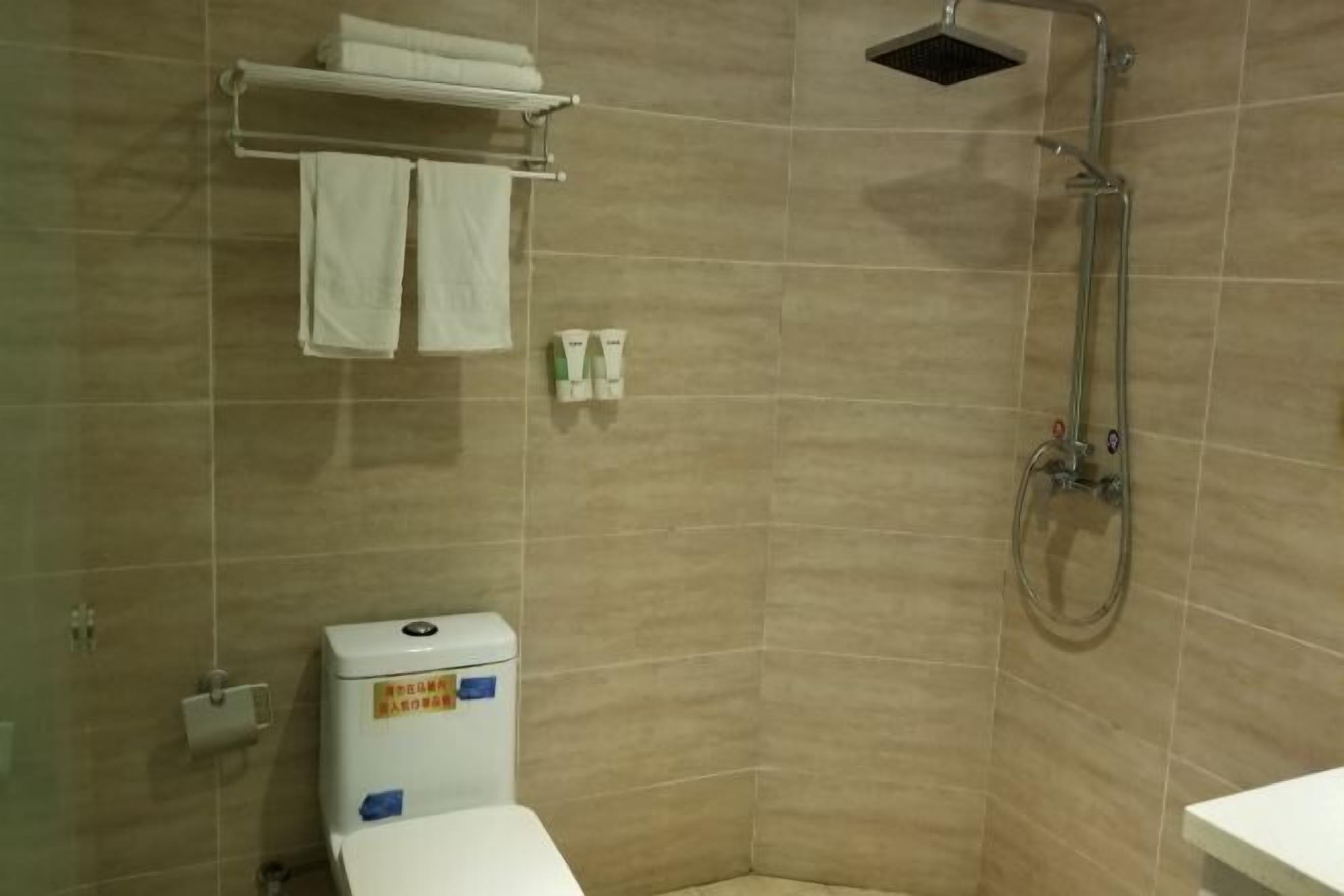 Un baño con baldosas color beige, un inodoro blanco, una ducha y un perchero con dos toallas.