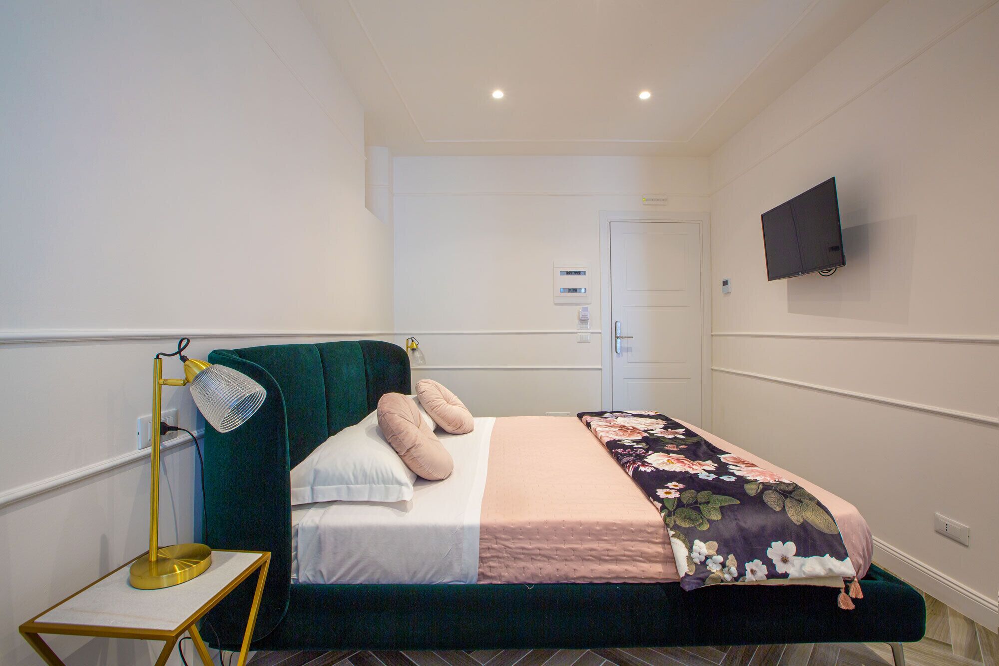A bedroom with a bed, a bedside lamp, a TV on the wall, and a door in the background.