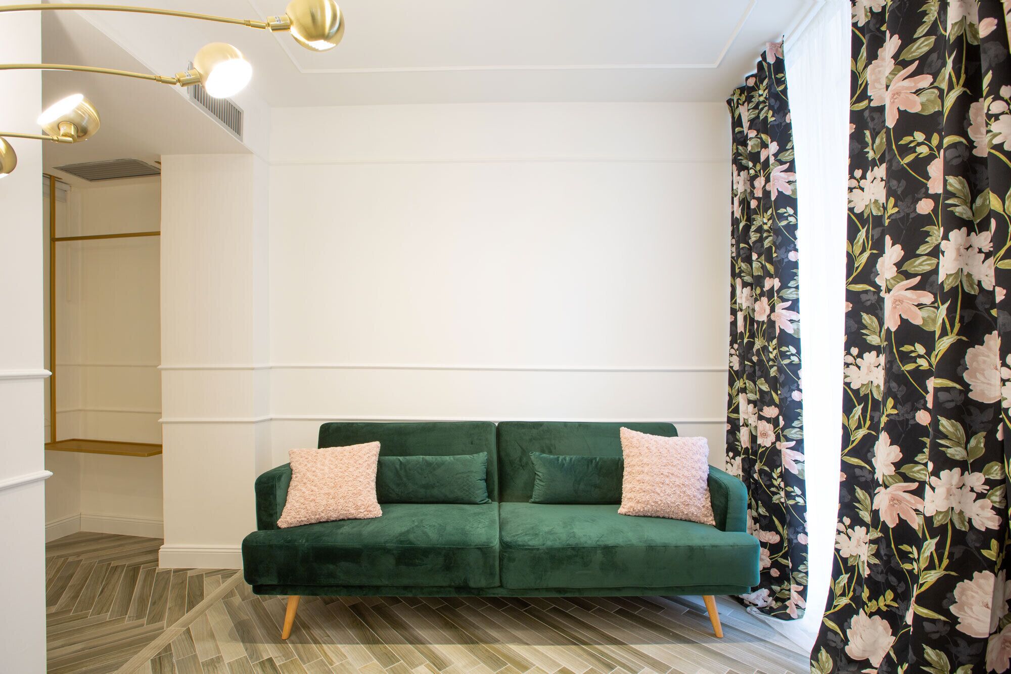 Ein grünes Samtsofa mit zwei rosafarbenen Kissen, Holzboden und einem blumigen Vorhang.