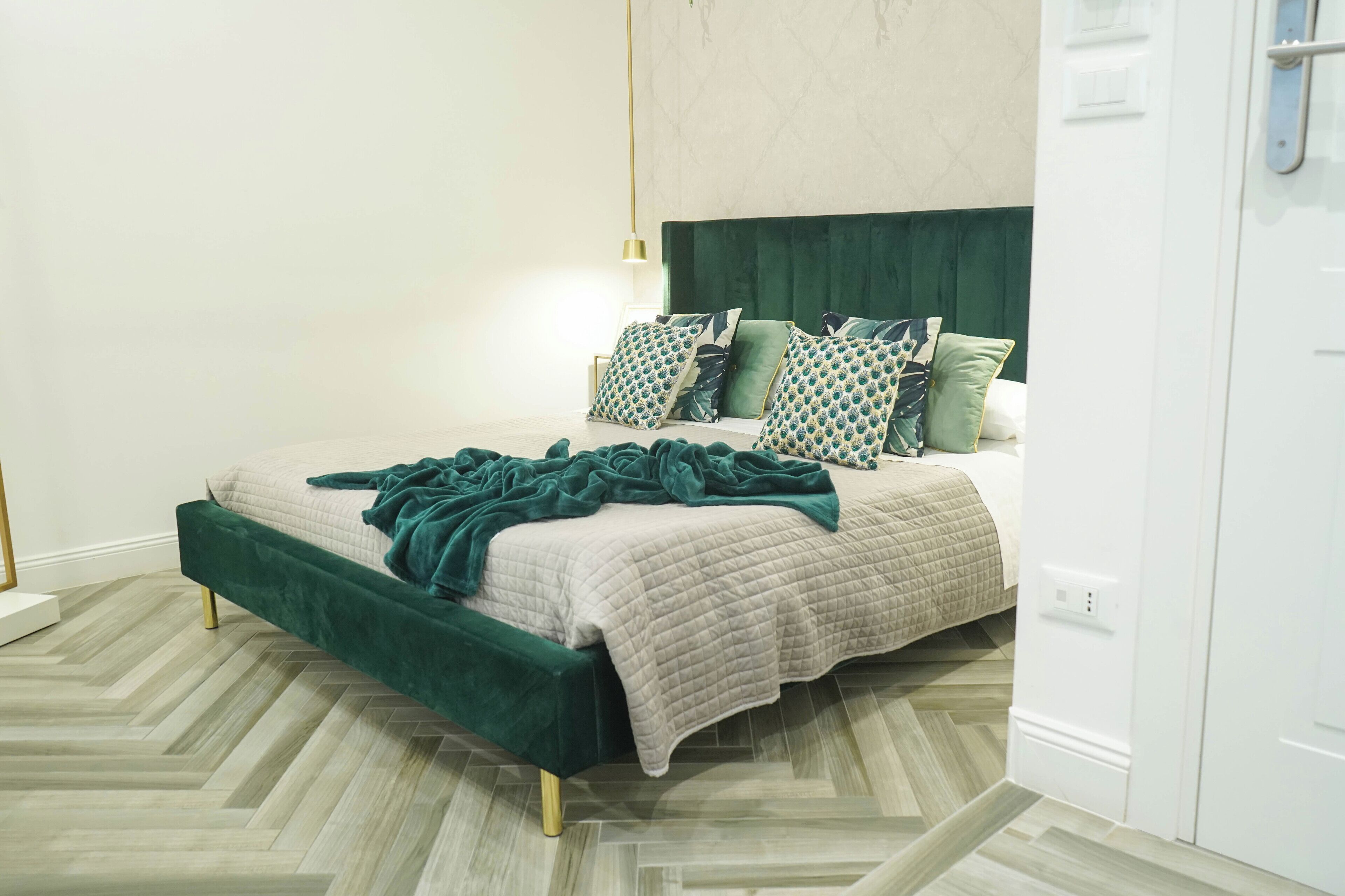 Un dormitorio moderno con una cama tapizada en verde, almohadas estampadas y un suelo de madera con patrón de espiga.