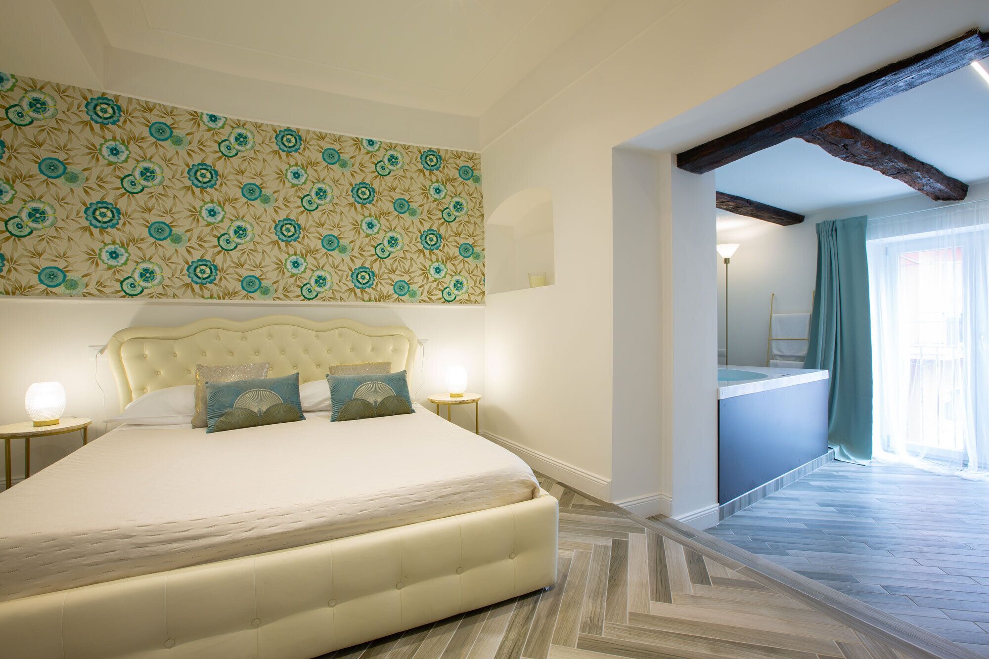 Un dormitorio con una cama grande, dos mesitas de noche, un papel tapiz estampado y vistas a un baño a través de un vano abierto.