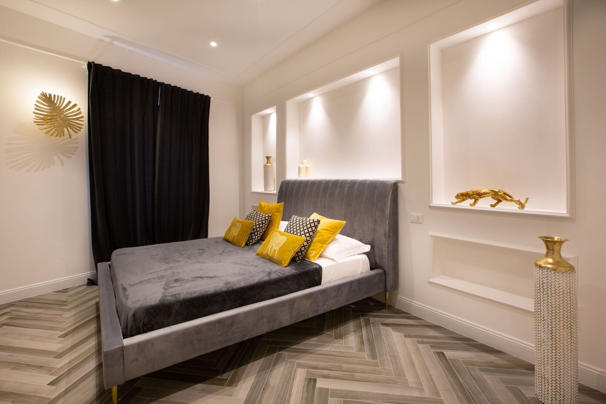 A modern bedroom with a grey upholstered bed, a herringbone-patterned floor, and a decorative mirror above the bed.