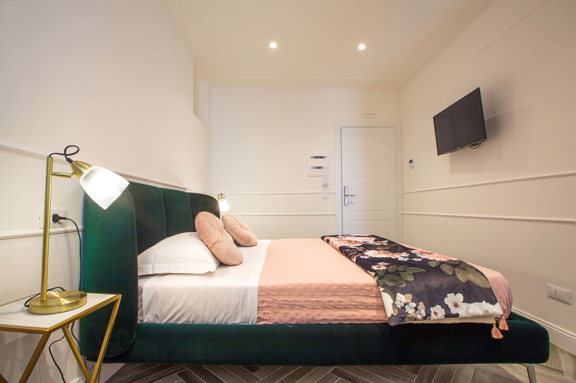 Un dormitorio ordenado con cama, lámpara de noche, televisor montado en la pared y una puerta al fondo.