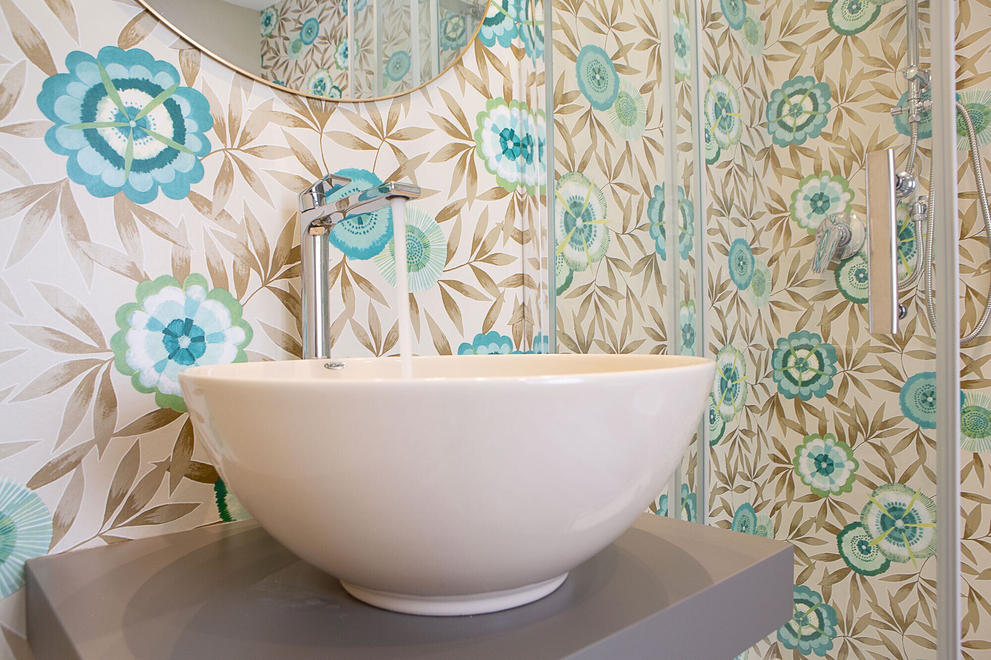 Ein Badezimmer mit einem großen, weißen Waschtischbecken, einer Dusche mit Glastür und blumigem Wandpapier.