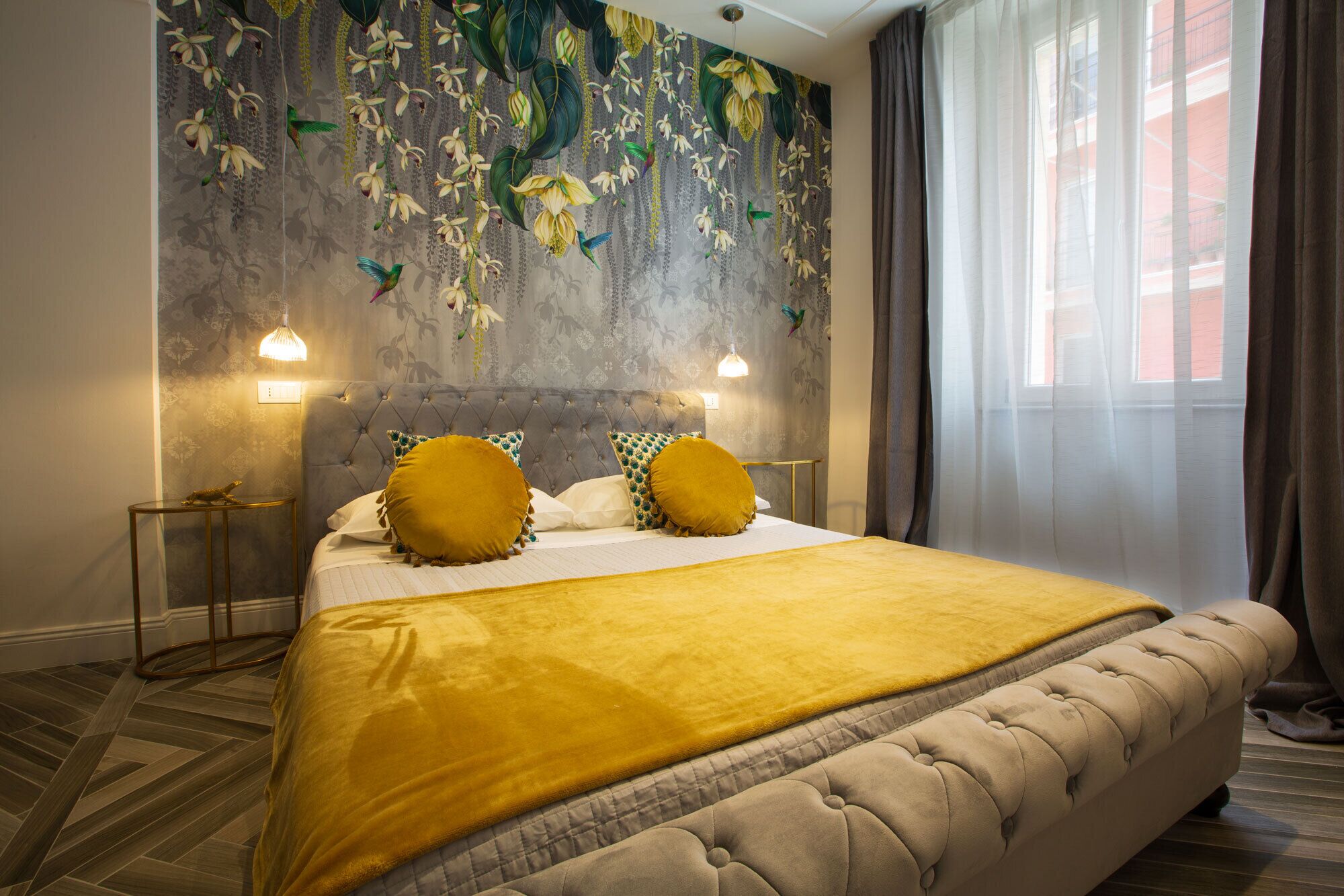 A bedroom with a tufted headboard, a large bed with a yellow blanket, two matching pillows, and a decorative wall with hanging plants and birds.