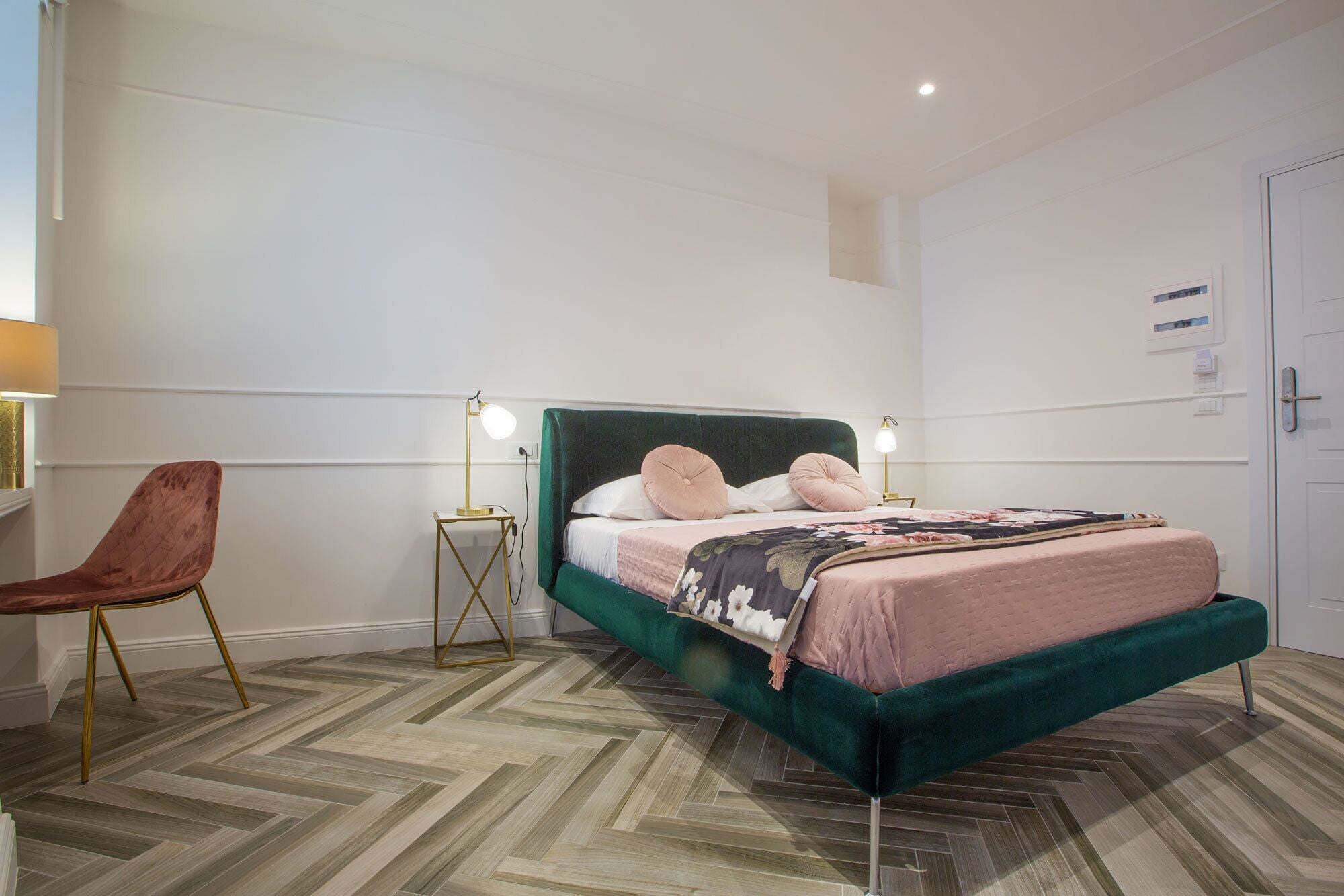 A bedroom with a green upholstered bed, two bedside lamps, a wooden herringbone floor, and a red chair.