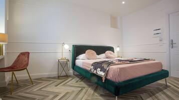 A bedroom with a green upholstered bed, two bedside lamps, a wooden herringbone floor, and a red chair.