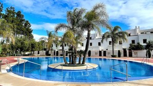 Una piscina con palmeras, un suelo de ladrillo rojo y un edificio blanco al fondo.