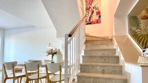 Una escalera con acabado de mármol, un comedor con sillas blancas y una mesa, y un cuadro abstracto de gran tamaño en la pared.