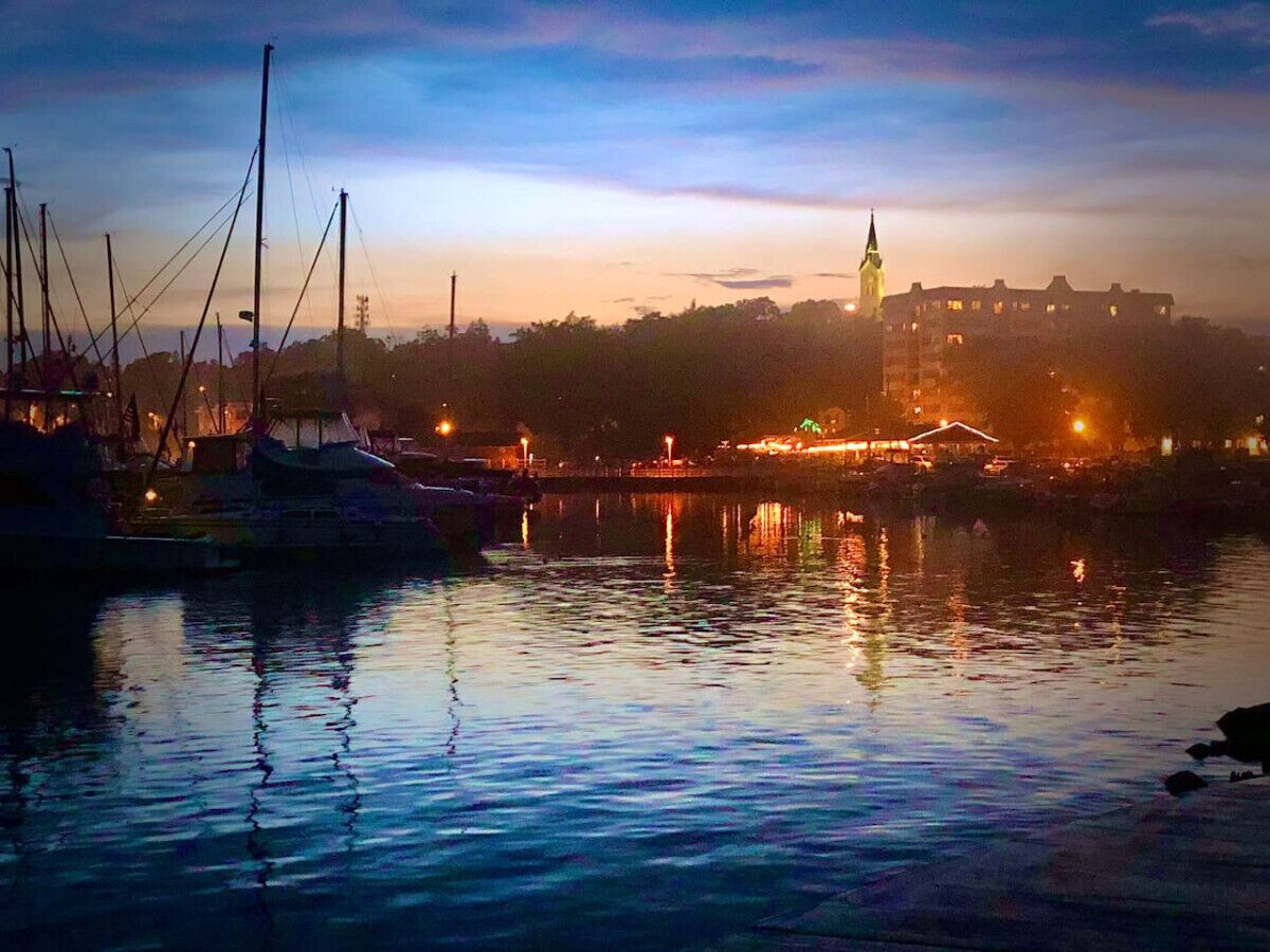 A marina at dusk with boats docked, a prominent building with a spire in the background, and calm water reflecting the lights.