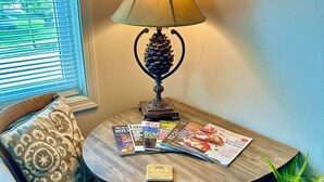 Un coin salon avec des sièges, une table, une lampe, des magazines, un petit livre et une plante en pot.