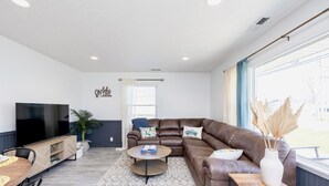 A modern living room with a brown leather sofa, a round coffee table, a flat-screen TV, and a large window with curtains. - Island Cottage w/ Pool Table & Walking distance to Cranberry!  (Huntsville)
