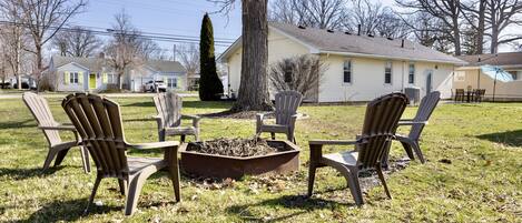 A backyard with Adirondack chairs around a fire pit, a large tree, and houses in the background.