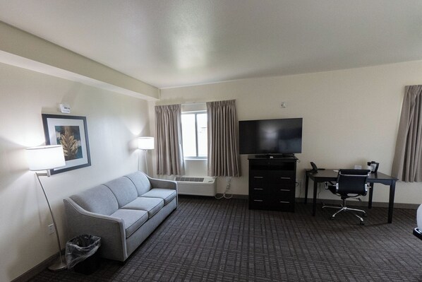 A hotel room with a grey sofa, a desk with a chair, a television, a framed picture on the wall, and a window with curtains.