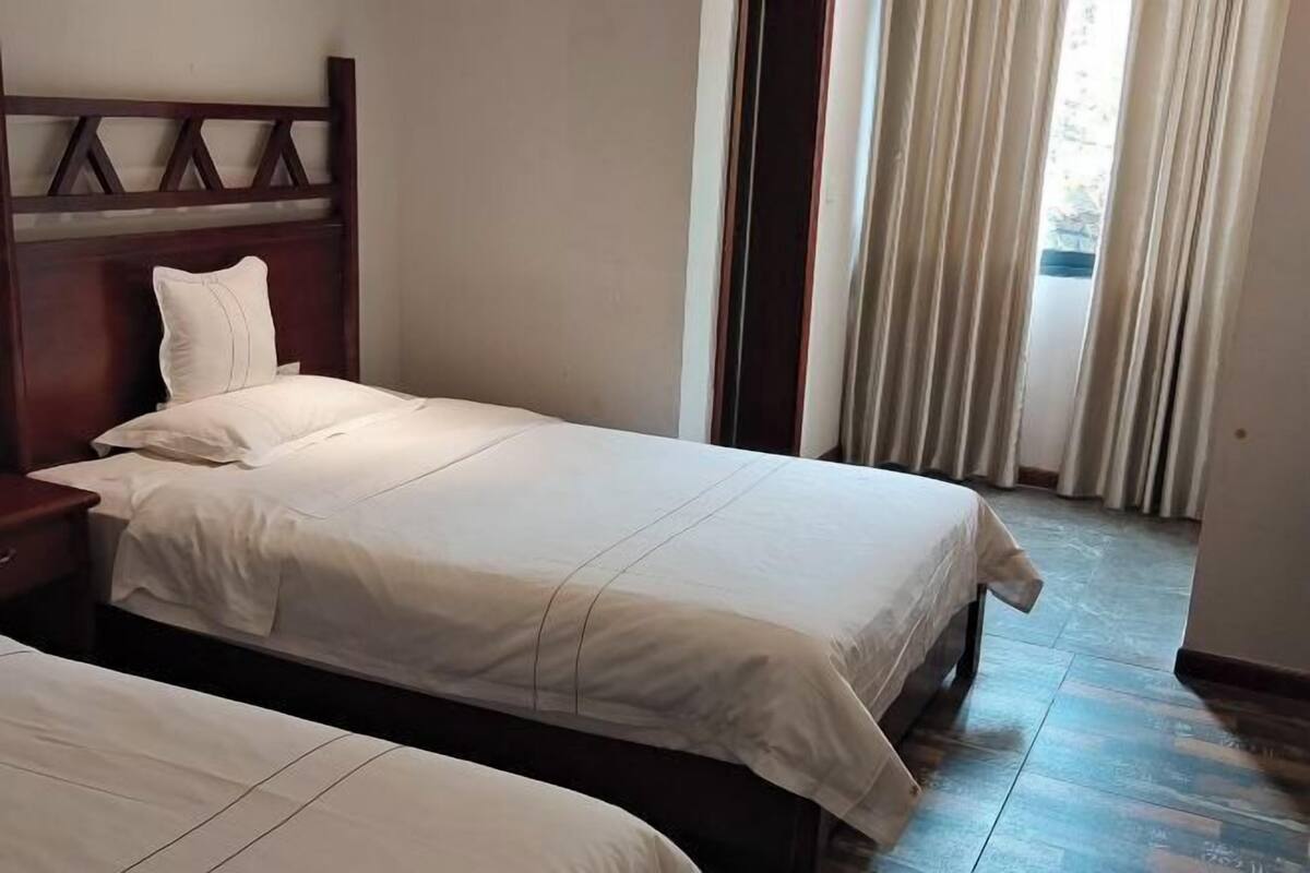 A hotel room with two beds, a wooden headboard, a nightstand, and a view of the outdoors through a window with curtains.