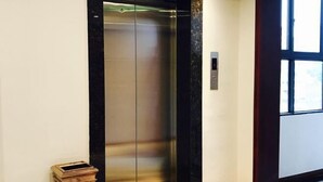 An elevator with a metallic finish and a marble-like base, adjacent to a wall with a wooden trim and a small trash bin. - Nanyi Hotel (Chishui)