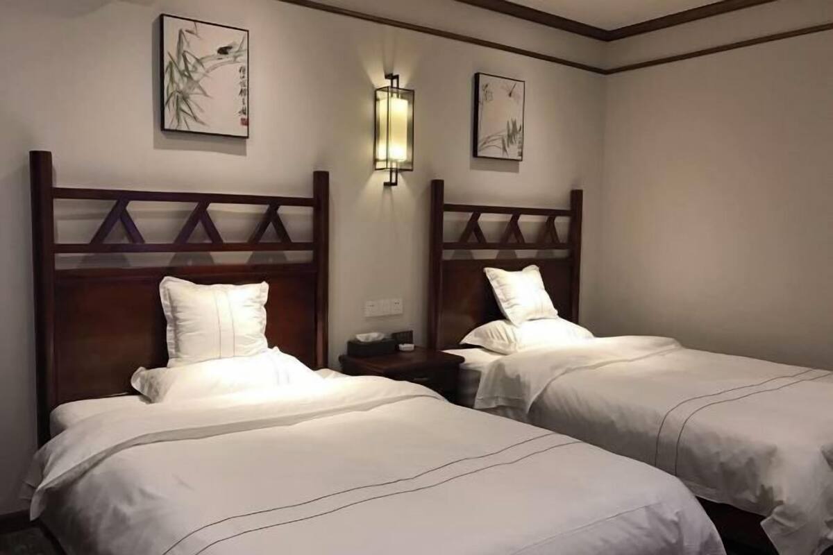A hotel room with two beds, wooden headboards, white bedding, wall-mounted light fixtures, and framed artwork on the walls.
